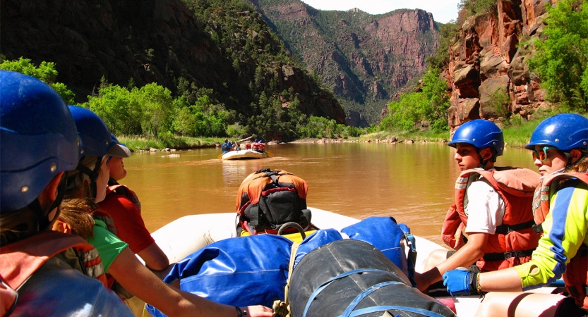 From the back of a raft, students are seen in front wearing safety gear. The water is calm, and there is another raft in the distance. The river is framed by tall canyon walls.