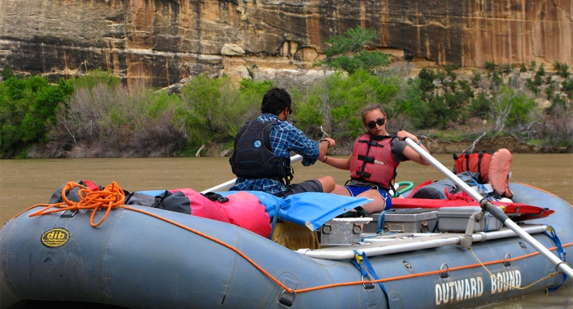 two people paddle a raft on calm water. behind them are tall canyon walls.