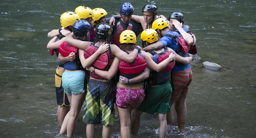a group of people wearing safety gear stand in ankle-deep water, huddled in a circle, with their arms around each other.
