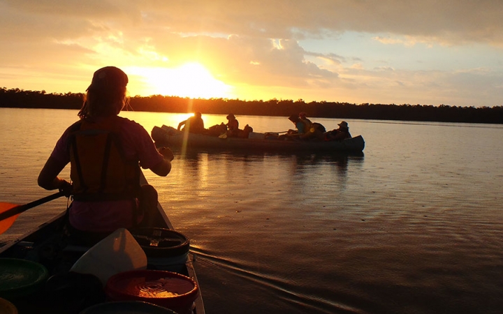 the silhouettes of people paddling canoes are illuminated as the sun sets behind trees on the horizon. The water is calm.
