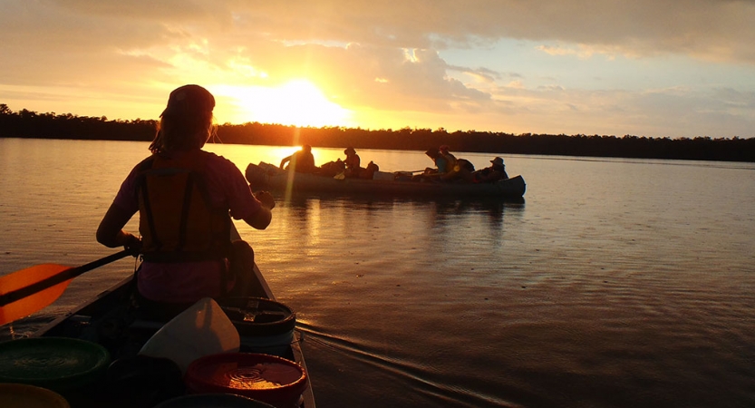 the silhouettes of people paddling canoes are illuminated as the sun sets behind trees on the horizon. The water is calm.