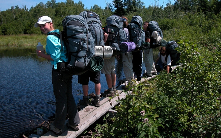 A group of students wearing backpacks stand on a narrow wooden plank over a body of water. Thick greenery surrounds the water. 