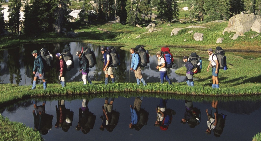 A group of people wearing backpacks hike on a grassy strip of land between two small bodies of water. Their images are reflected in the body of water in the foreground.