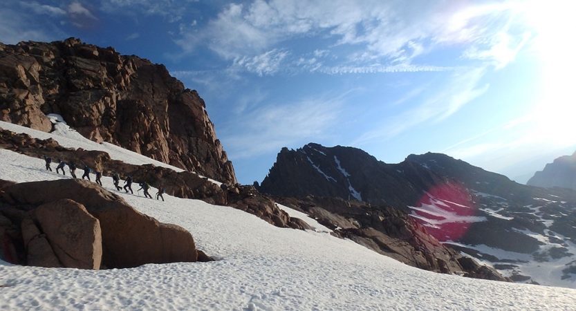 A line of people hike up a snowy incline in a mountainous landscape under blue skies.
