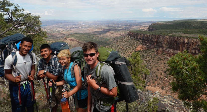 Four people wearing backpacks smile for a photo on an overlook high above a desert landscape.