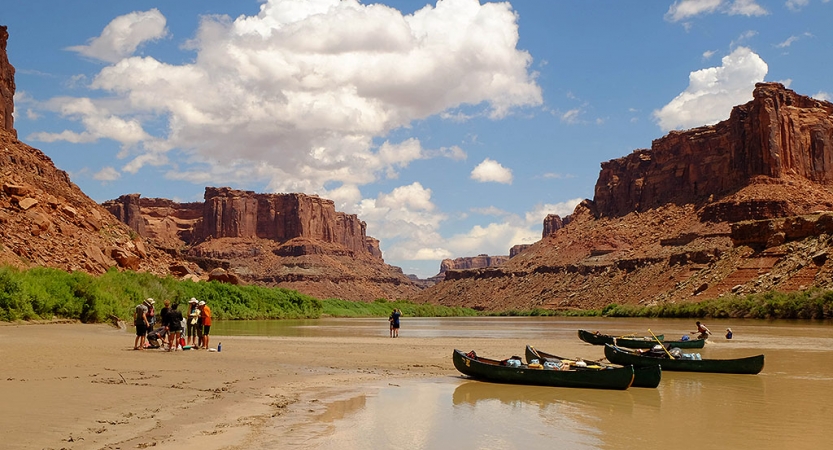 Three canoes rest on the shore of a river as a group of people stand nearby. There are tall, read mesas behind them.