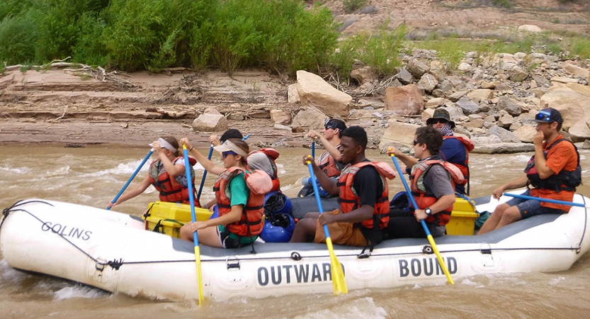 A group of students wearing safety gear paddle a green raft on a river. 