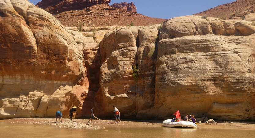 A raft is beached on a sandy shore, framed by tall canyon walls. There are people standing on the shore, too. 