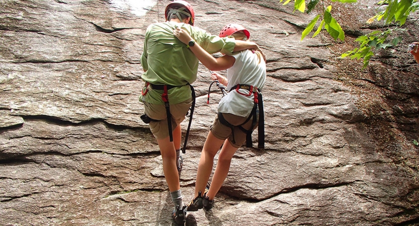 A parent and child wrap their arms around each other as they rappel down a rock wall. Both are wearing safety gear and are secured by ropes.