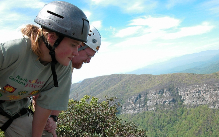 A parent and child wearing helmets look down over a vast mountainous landscape.