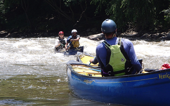 Two students paddle a canoe through whitewater, while another person who is likely an instructor watches them from another canoe.
