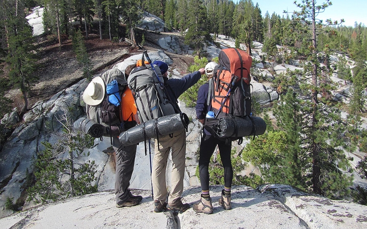 three people wearing backpacks stand on a rock while one of them points outward
