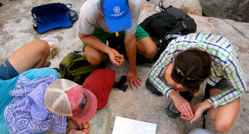 Three people snack on trail mix while examining a map spread out on a rock