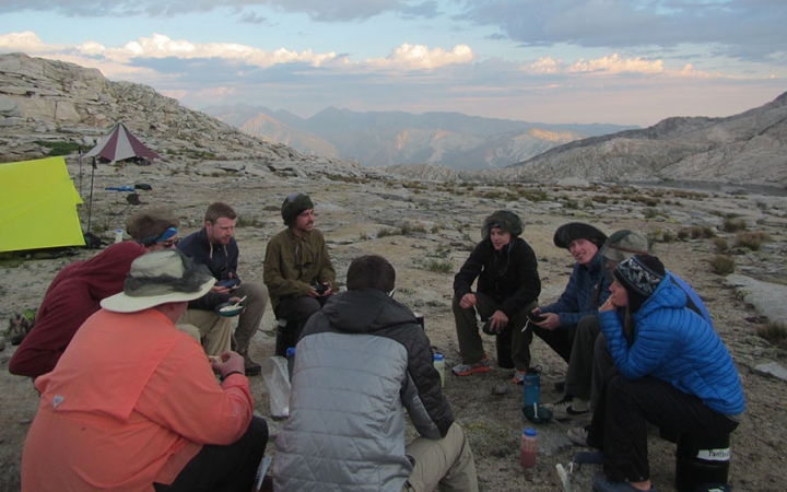 A group of outward bound students sit in a circle amidst a rocky landscape. Their tents sit nearby. There are mountains in the background. 