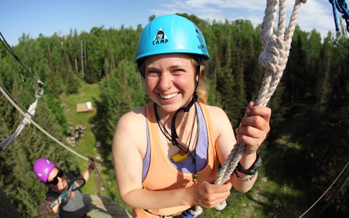 A person wearing safety gear and secured by ropes smiles as they make their way through a high ropes course.